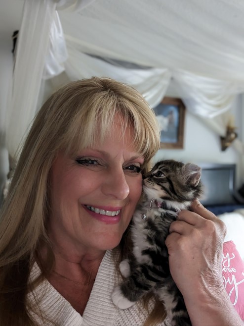 An image of a smiling woman with long blonde hair. She has a small kitten perched on her shoulder, looking content. The kitten is striped gray, with white paws, a white chin, and a white chest.