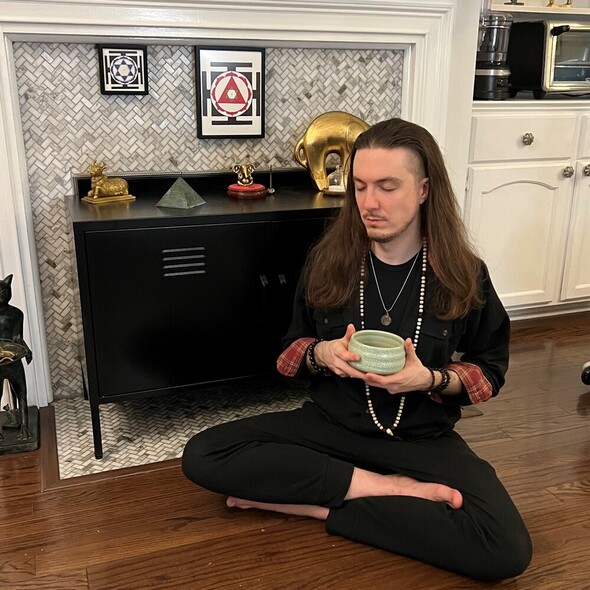 An image of Psychic Tobias, a young man with long brown hair. He is sitting cross-legged on the floor with a bowl in his hands. seeming meditative.